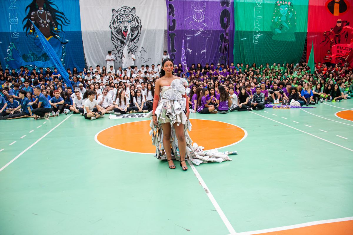 Aluna do Alcina desfilando com vestido de papel jornal reciclado no Dia D 2025, tendo as bandeiras das 6 equipes ao fundo